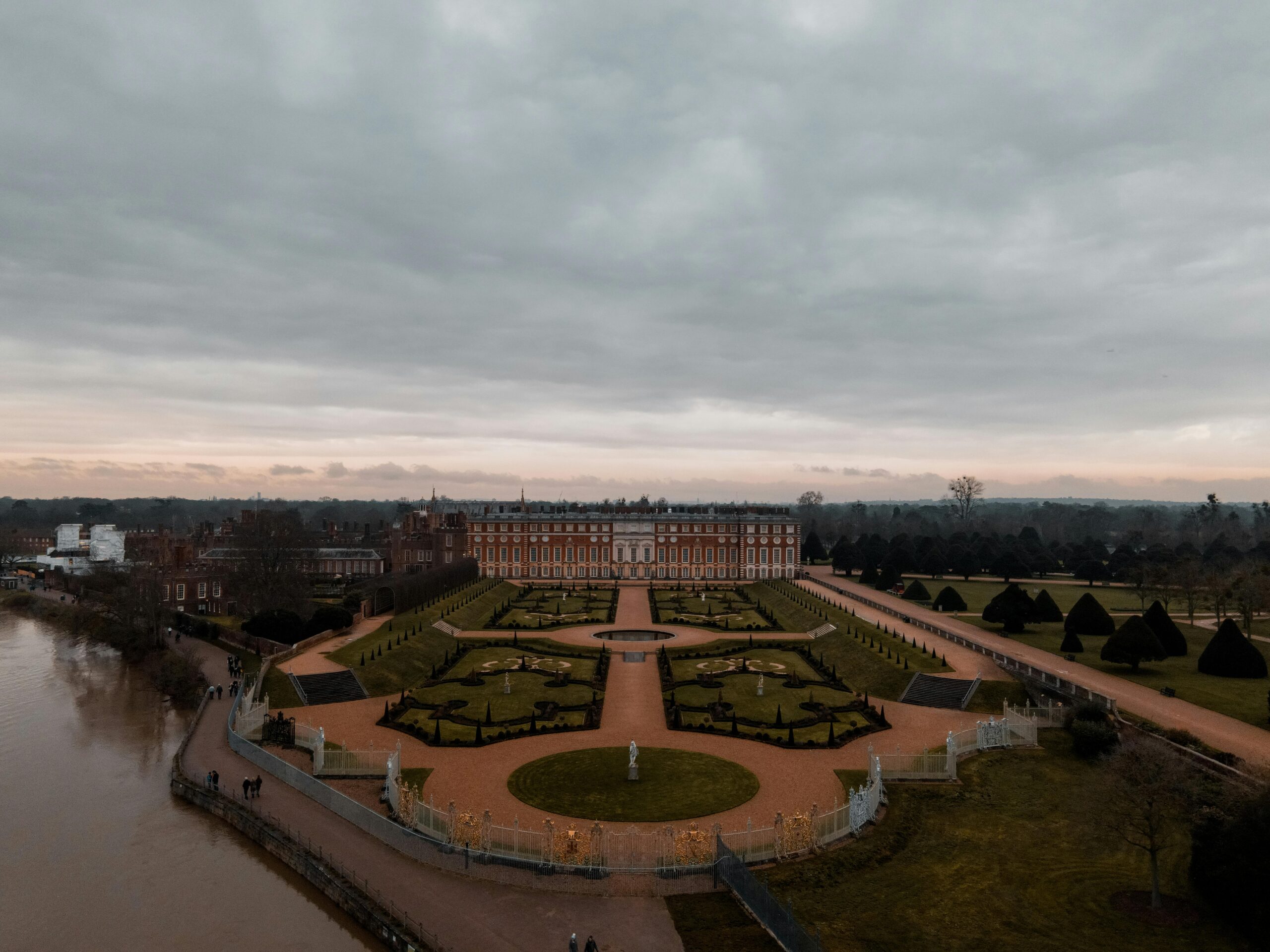 Drone view of historic Hampton Court Palace and gardens on a cloudy day, showcasing its grandeur and serene beauty.